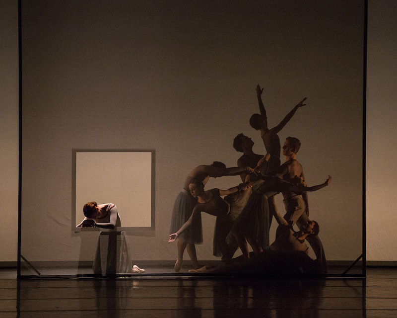 Dancers in an intricate tableau behind a square scrim. One dancer knells apart from the group. 